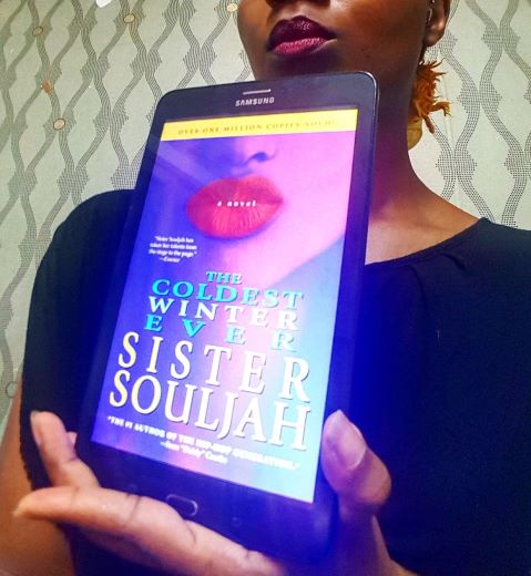 A Black woman(me) is partly visible, holding a tablet displaying the cover of The Coldest Winter Ever. The book cover shows a Black girl's face from the nose down, her full lips painted bright pink. The woman holding the book is also only visible from nose down and wearing a similar lipstick