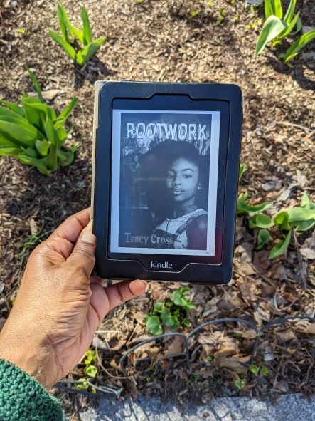 The ebook cover of Rootwork held above a tender new spring garden. The cover depicts a young black girl with soft lush hair, smiling enigmatically at the reader.