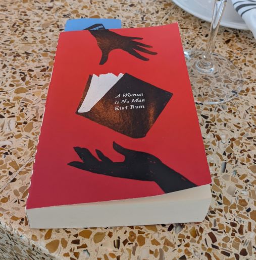 A photo of the paperback classic edition of A Woman Is No Man. The cover is black and red and depicts the a silhouetted hand dropping a book into another hand.