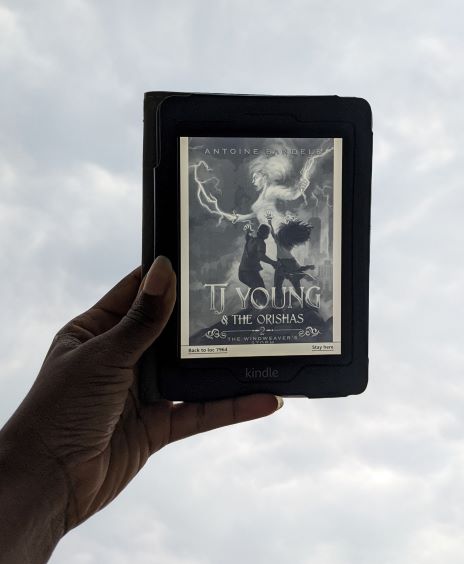 A Black woman's hand(my hand) holds up the Kindle cover of The Windweaver's Storm up against the cloudy blue and white sky. The cover shows a teenaged black boy and girl pointing at a giant female figure made of lightning and fire in the distance.