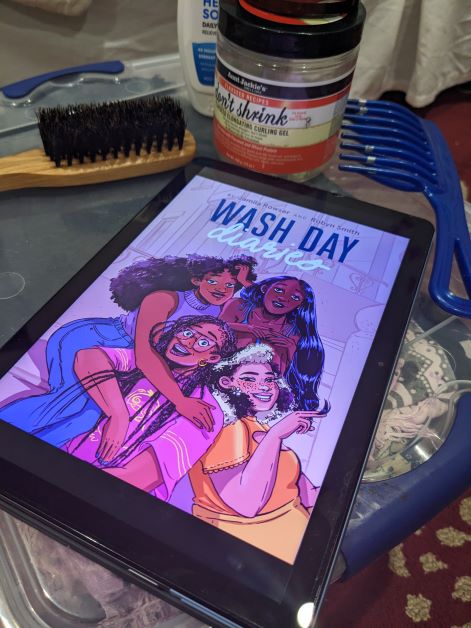 The purple cover of Wash Day Diaries, depicting a diverse foursome of Black women smiling, posing and embracing, lays on a table surrounded by hair gel, a boar bristle brush, a big tooth comb, and other Black hair tools